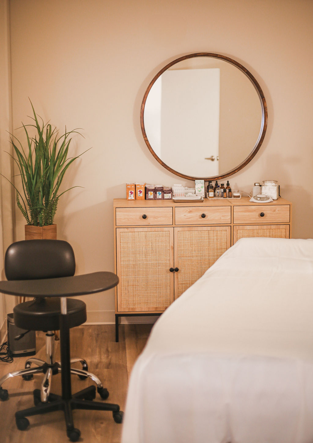 Acupuncture treatment room featuring a white treatment table, practitioner's stool, woven cabinet with supplies, round mirror, and green plant